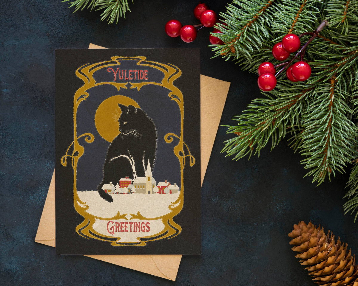 A square holiday greeting card with a vintage-style illustration of a black cat in front of a yellow moon and small houses, placed on top of a kraft envelope. The card is styled on a dark textured surface with evergreen branches, red berries, and a pinecone around it.