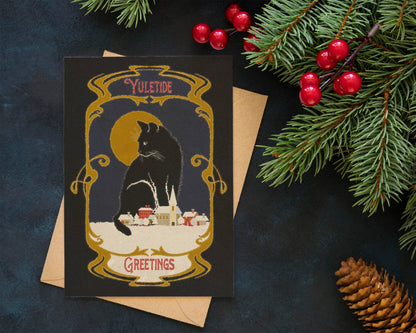 A square holiday greeting card with a vintage-style illustration of a black cat in front of a yellow moon and small houses, placed on top of a kraft envelope. The card is styled on a dark textured surface with evergreen branches, red berries, and a pinecone around it.
