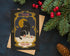 A square holiday greeting card with a vintage-style illustration of a black cat in front of a yellow moon and small houses, placed on top of a kraft envelope. The card is styled on a dark textured surface with evergreen branches, red berries, and a pinecone around it.