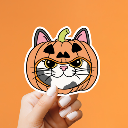 A hand with white manicured nails holds a die-cut sticker shaped like a pumpkin cat head. The sticker features an orange pumpkin body with a carved face, pink inner ears, and a small green stem, set against a solid orange background.