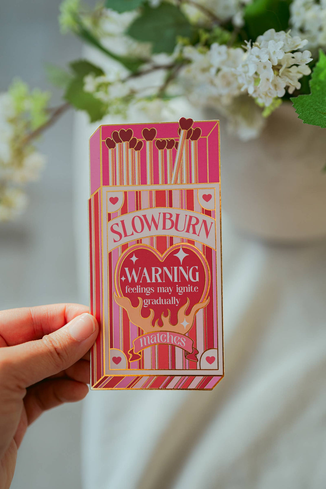 Bookmark of a box of Slowburn matches held by a hand with flowers in the background