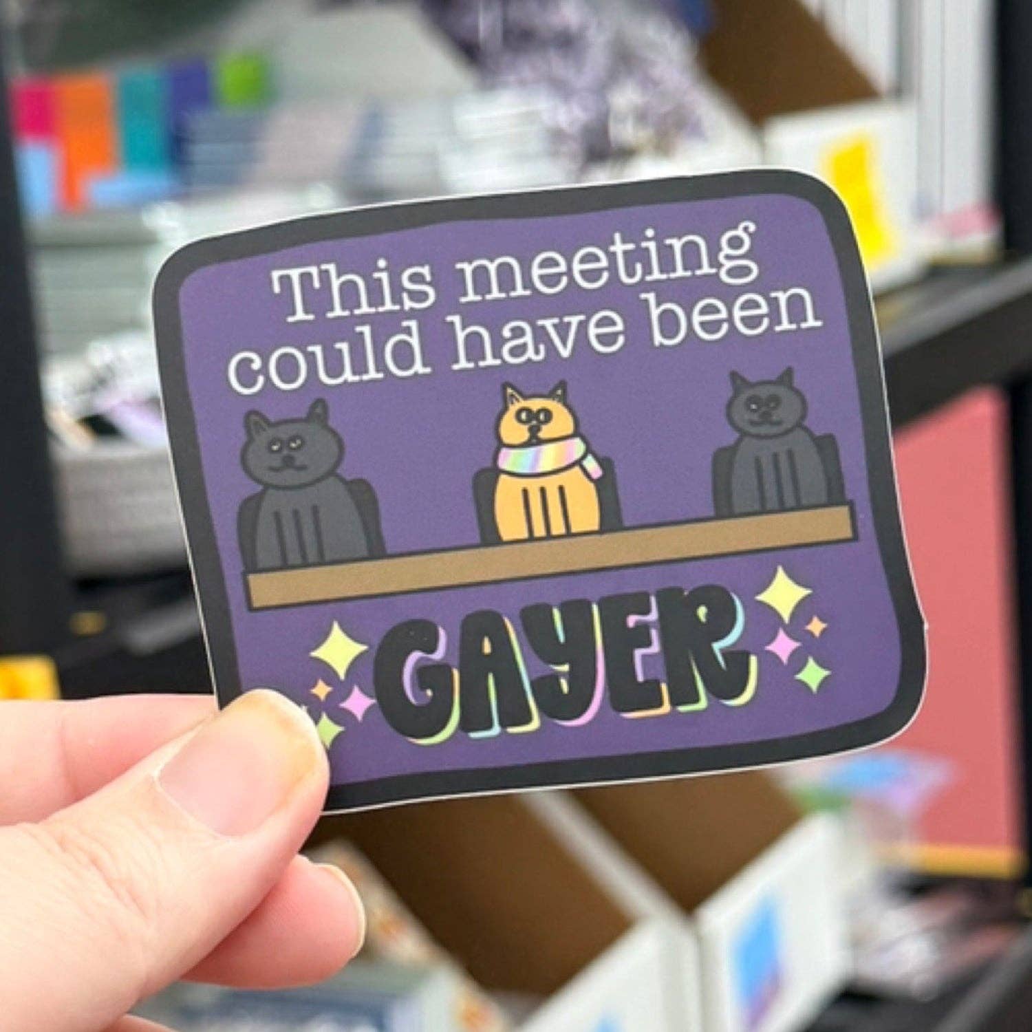 A hand holds a die-cut sticker with a purple background and cartoon cats sitting at a table. The sticker reads &