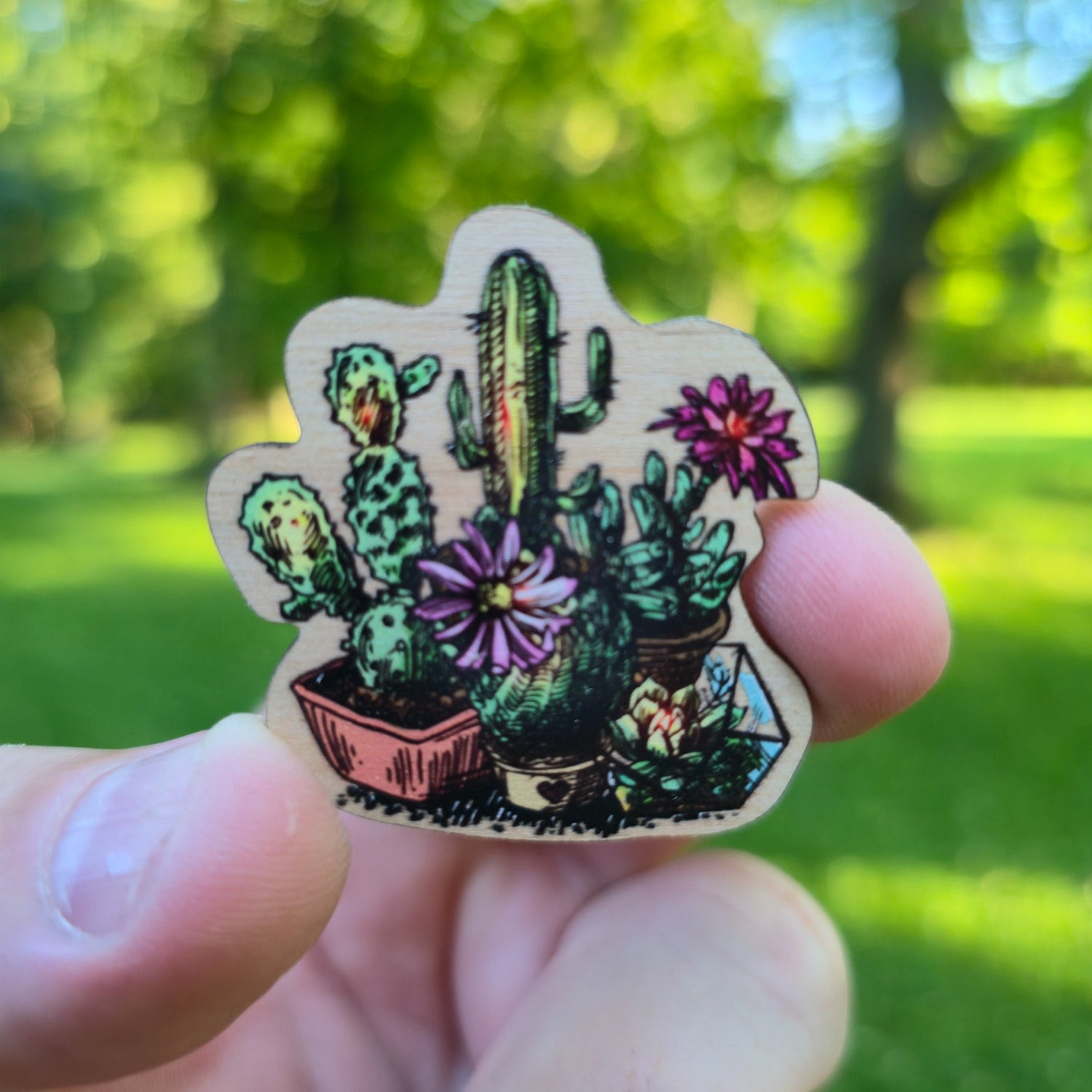 Magnet of potted cacti held in a hand with a green outdoor background