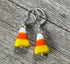 A pair of Halloween Theme Earrings shaped like candy corn, from Enlivenadorn by Gwynstone, featuring white, orange, and yellow stripes with silver hooks, displayed on weathered wood.