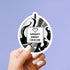 A hand with white manicured nails holds a round sticker or decal featuring a black, white, and gray swirled design with a central white box that reads &