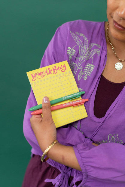 Person holding a yellow notepad with colorful pens against a green background