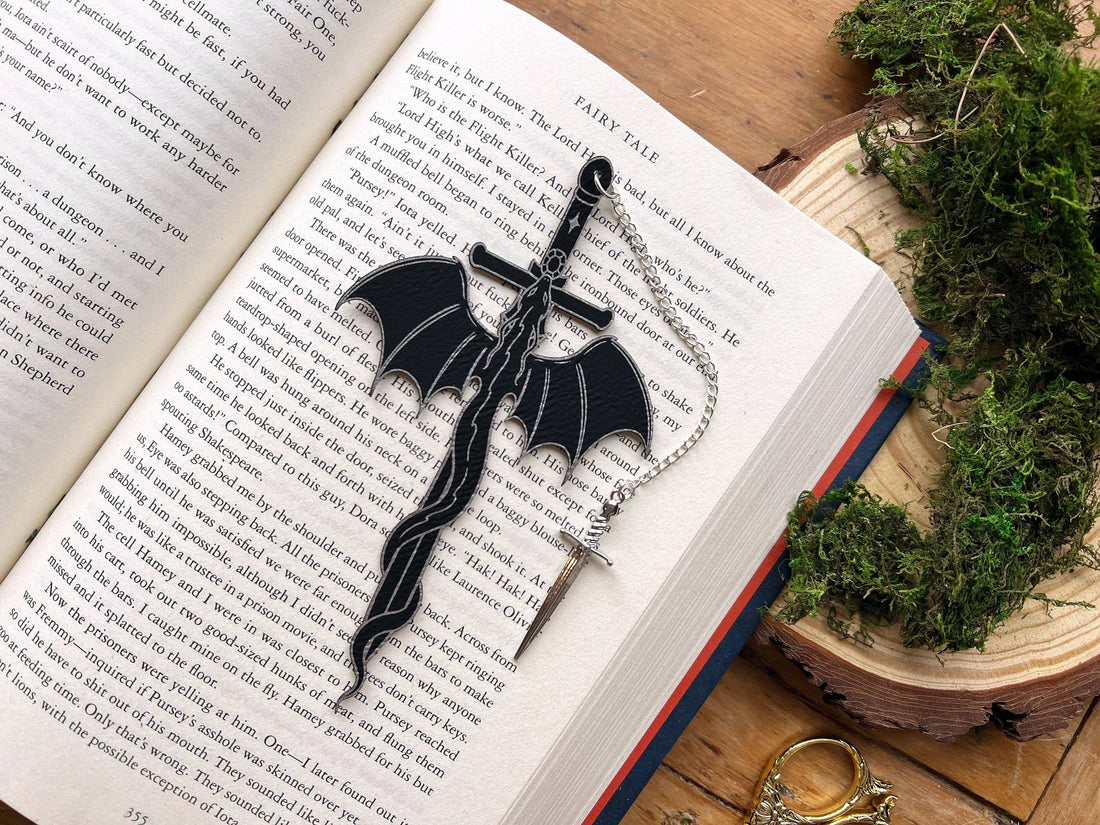 Black dragon-shaped bookmark on a silver chain on an open book with greenery around