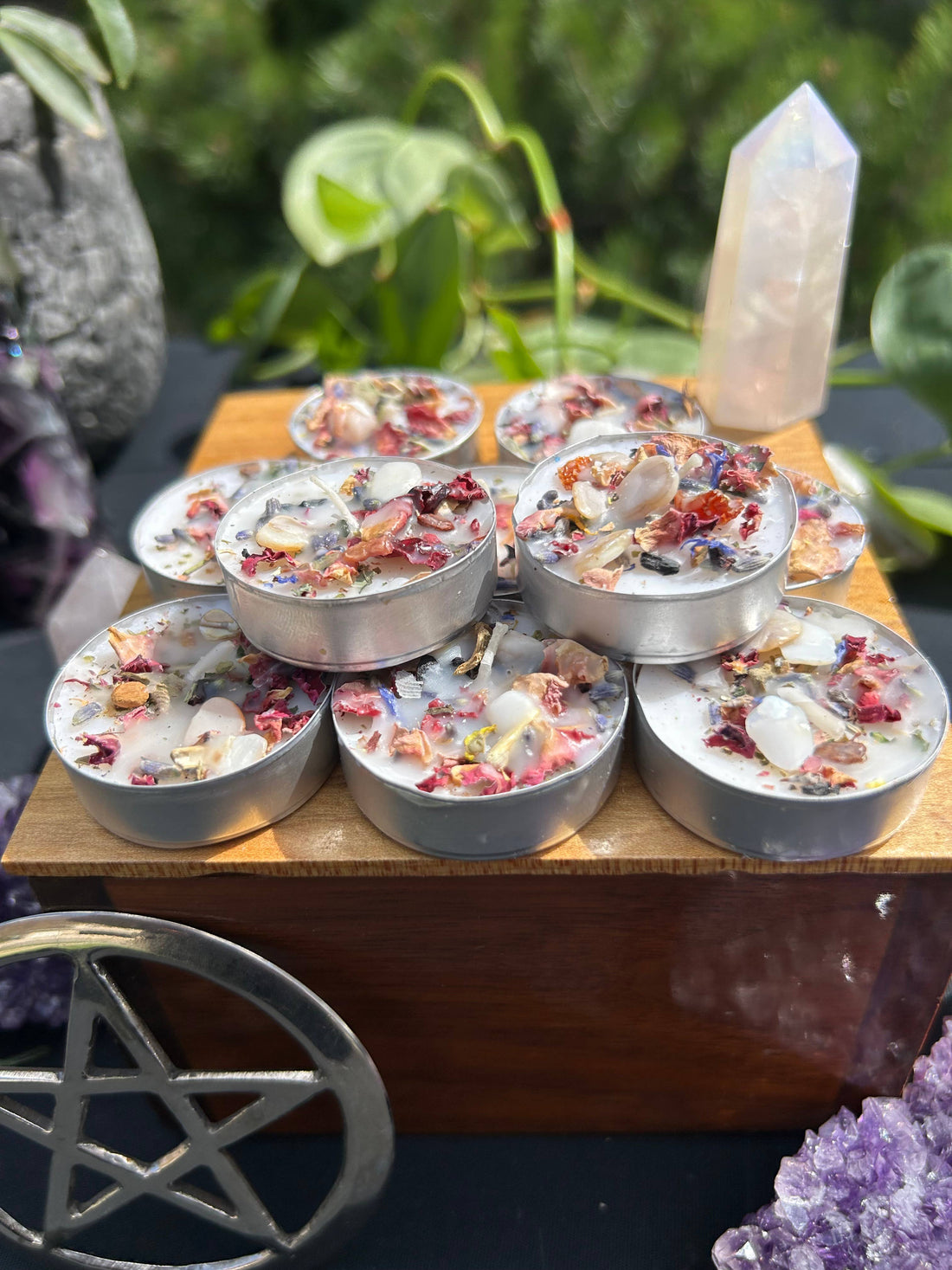 Small candles with herbs on a wooden surface, surrounded by greenery and crystals.
