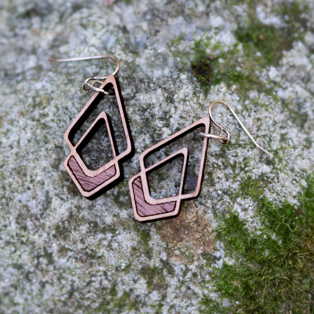 Two geometric drop earrings with layered diamond and triangle shapes, featuring a reddish-brown wood patterned inlay and a silver-tone metal border and fishhook wires, displayed on a mossy gray stone surface.