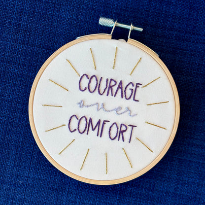 "Courage over comfort". "courage" and "comfort" are hand embroidered in dark purple embroidery thread and are all capital letters in a print handwriting font. "over" is stitched in light purple embroidery floss in a lowercase cursive handwriting font. Quote is centered in a burst of metallic gold rays. Fabric is white cotton. Framed in light wood embroidery hoop as wall art or Christmas ornament.