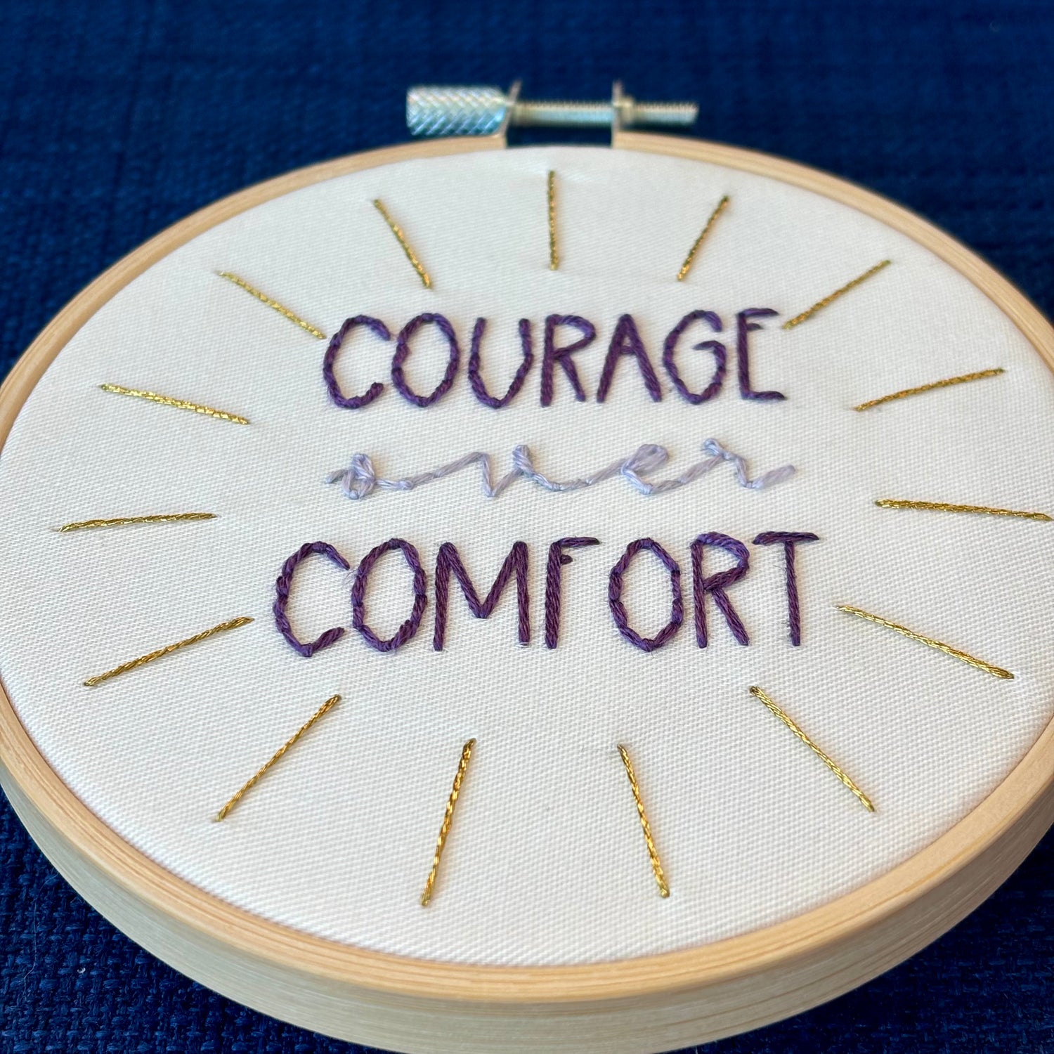 "Courage over comfort". "courage" and "comfort" are hand embroidered in dark purple embroidery thread and are all capital letters in a print handwriting font. "over" is stitched in light purple embroidery floss in a lowercase cursive handwriting font. Quote is centered in a burst of metallic gold rays. Fabric is white cotton. Framed in light wood embroidery hoop as wall art or Christmas ornament.