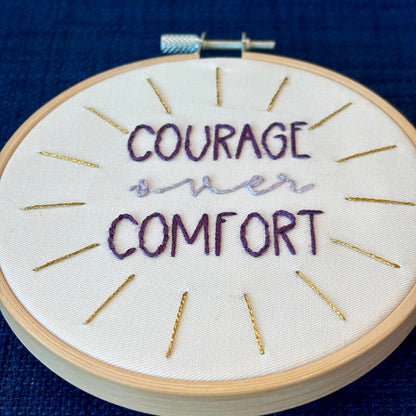 "Courage over comfort". "courage" and "comfort" are hand embroidered in dark purple embroidery thread and are all capital letters in a print handwriting font. "over" is stitched in light purple embroidery floss in a lowercase cursive handwriting font. Quote is centered in a burst of metallic gold rays. Fabric is white cotton. Framed in light wood embroidery hoop as wall art or Christmas ornament.