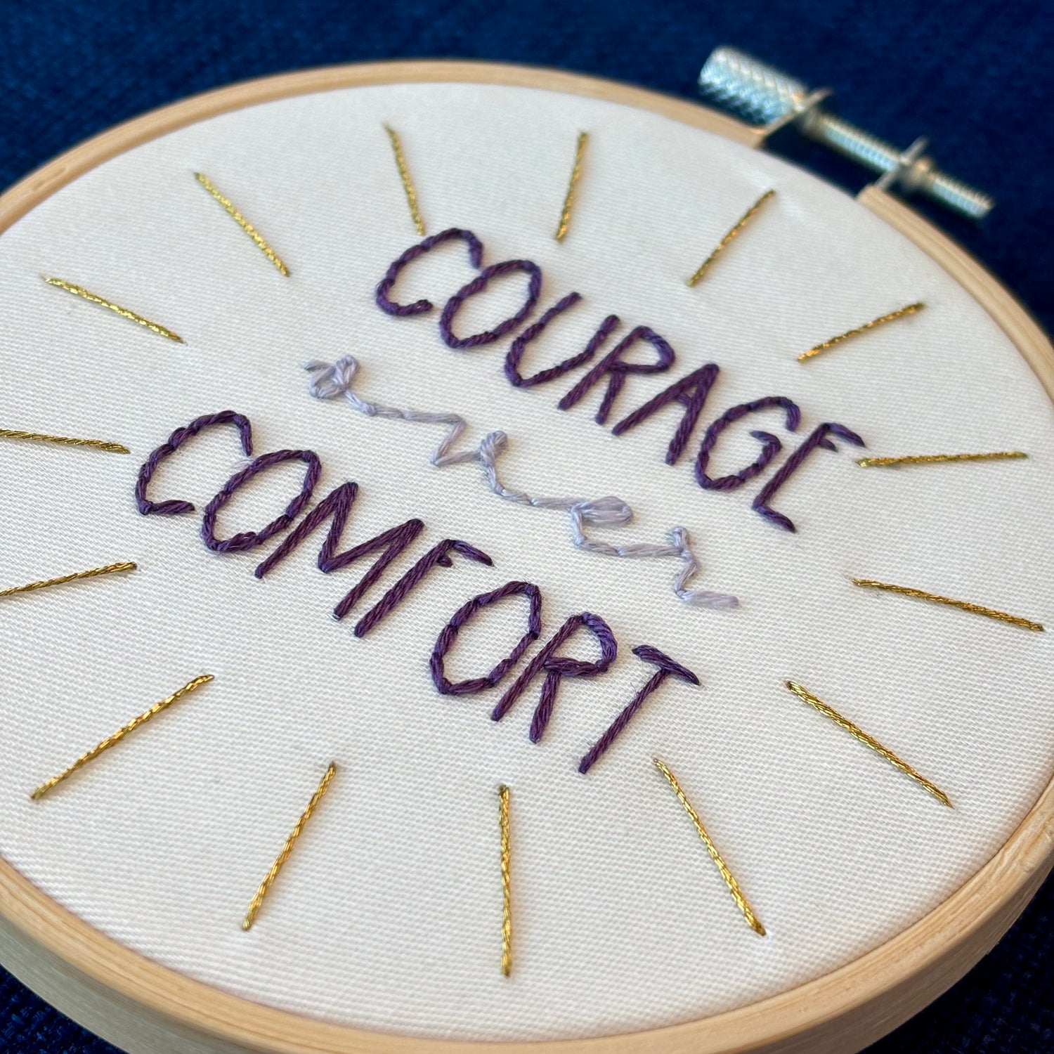 "Courage over comfort". "courage" and "comfort" are hand embroidered in dark purple embroidery thread and are all capital letters in a print handwriting font. "over" is stitched in light purple embroidery floss in a lowercase cursive handwriting font. Quote is centered in a burst of metallic gold rays. Fabric is white cotton. Framed in light wood embroidery hoop as wall art or Christmas ornament.