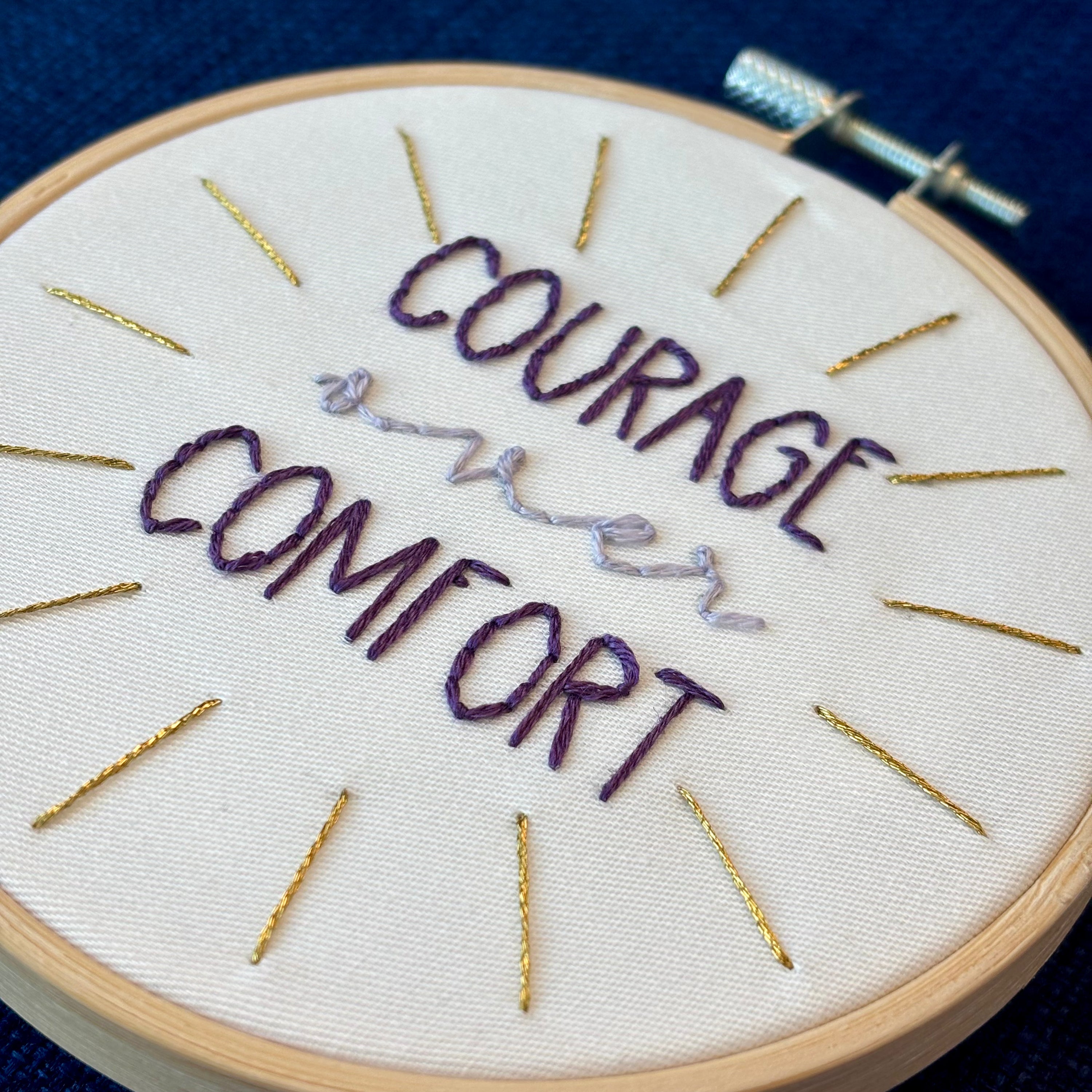 "Courage over comfort". "courage" and "comfort" are hand embroidered in dark purple embroidery thread and are all capital letters in a print handwriting font. "over" is stitched in light purple embroidery floss in a lowercase cursive handwriting font. Quote is centered in a burst of metallic gold rays. Fabric is white cotton. Framed in light wood embroidery hoop as wall art or Christmas ornament.