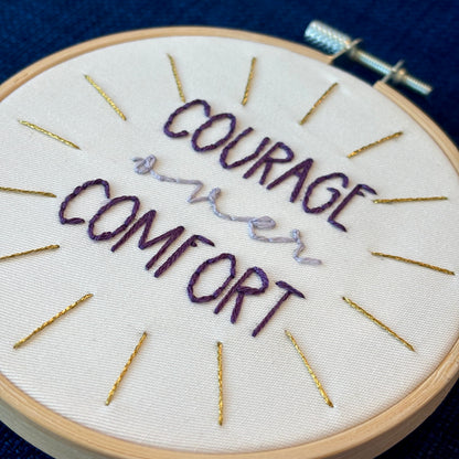 "Courage over comfort". "courage" and "comfort" are hand embroidered in dark purple embroidery thread and are all capital letters in a print handwriting font. "over" is stitched in light purple embroidery floss in a lowercase cursive handwriting font. Quote is centered in a burst of metallic gold rays. Fabric is white cotton. Framed in light wood embroidery hoop as wall art or Christmas ornament.