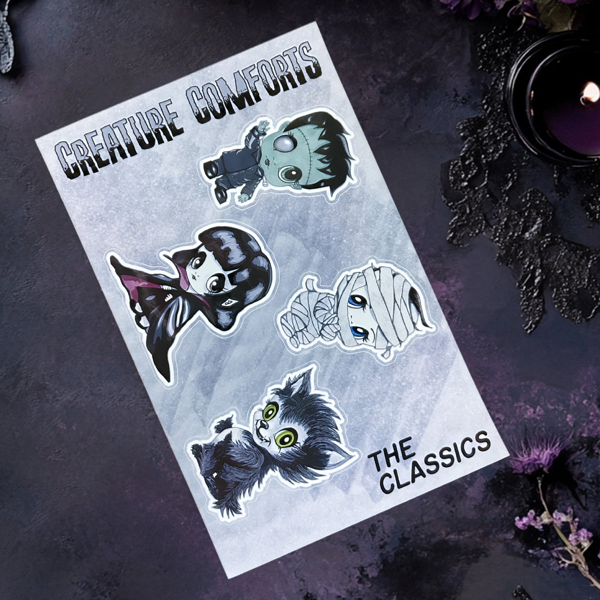 Creepy cute monster sticker sheet angled on dark purple stone with a black candle and dried purple flowers