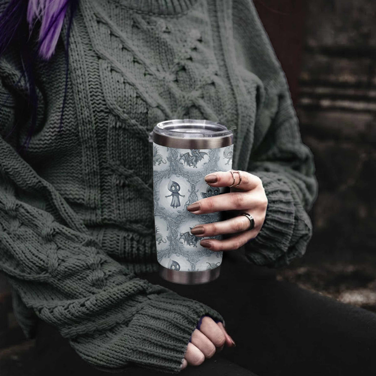 Woman with purple hair in a green cable-knit sweater holding the cool gray cryptid tumbler in a stone doorway
