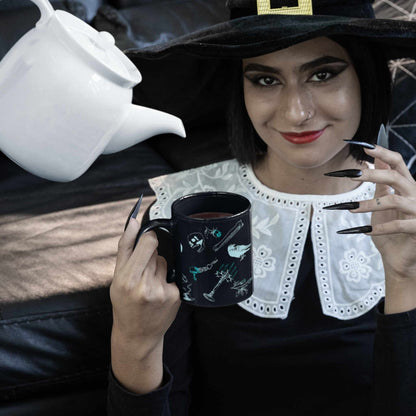 Person in a witch hat holding a cybergoth Victorian zombie icons mug while a white teapot pours