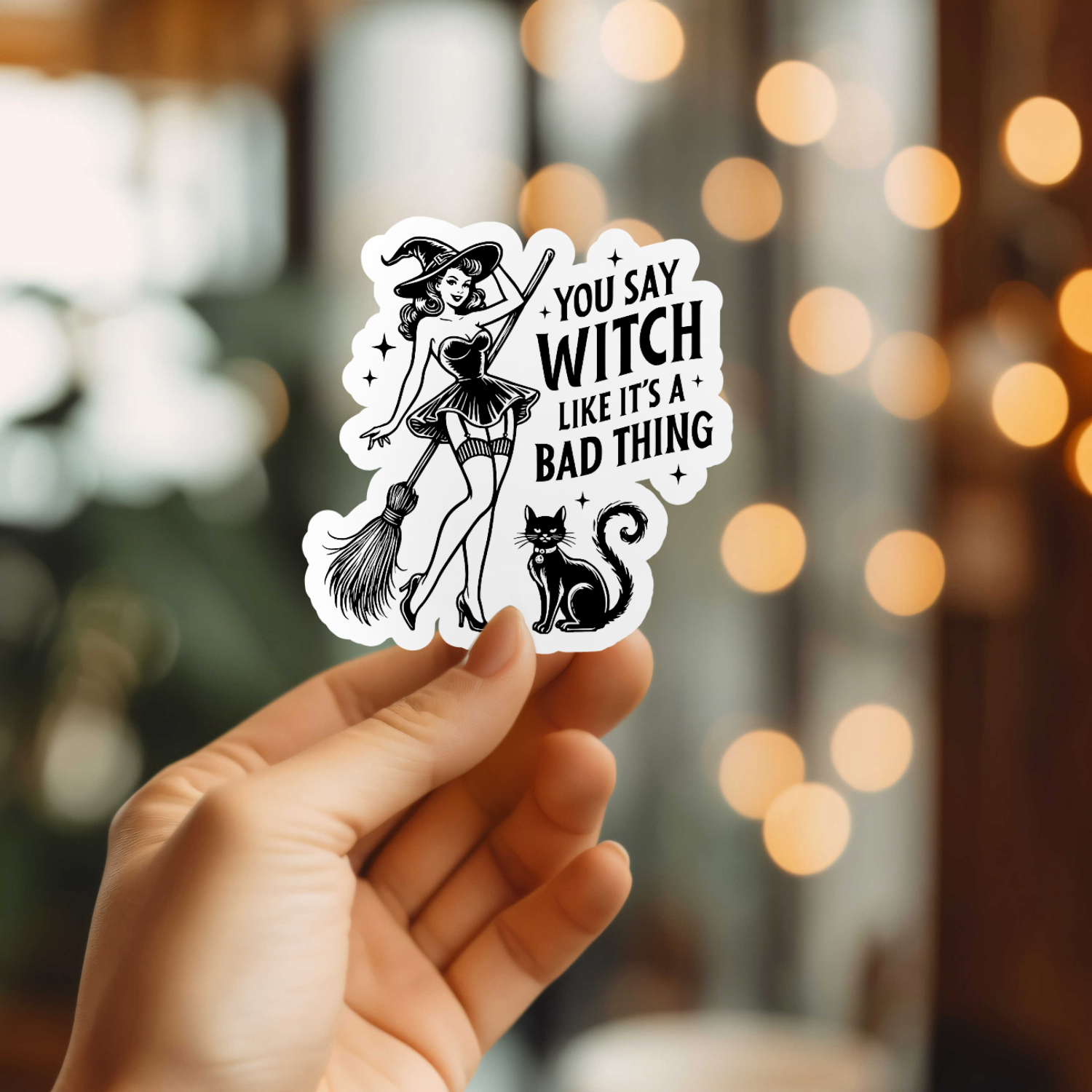 Hand holding a sticker with a witch design and text against a blurred bokeh background