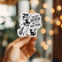 Hand holding a sticker with a witch design and text against a blurred bokeh background