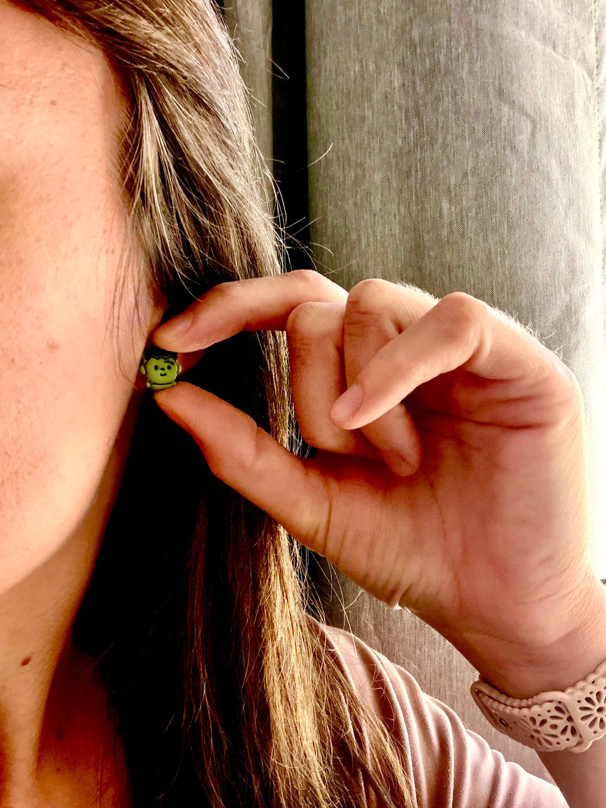A close-up lifestyle shot of a woman wearing a small green beaded earring, holding her hand near her ear to show the earring&