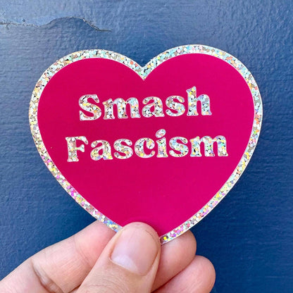 A hand holds a heart-shaped sticker with a bright pink background and a glittery multicolor border. The sticker features the text &