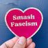A hand holds a heart-shaped sticker with a bright pink background and a glittery multicolor border. The sticker features the text &