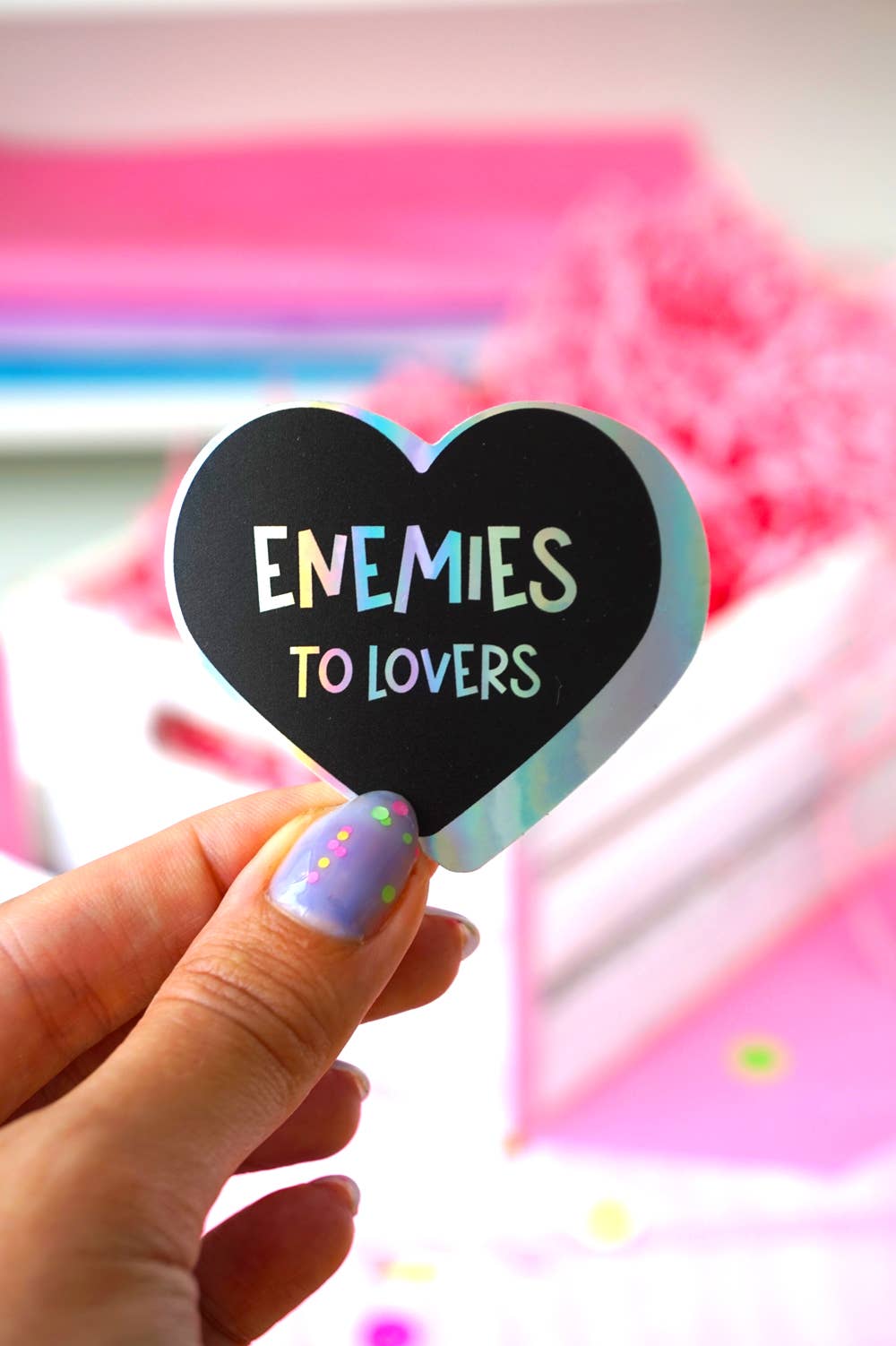 A hand with lavender glitter nail polish holds a heart-shaped black sticker that reads &