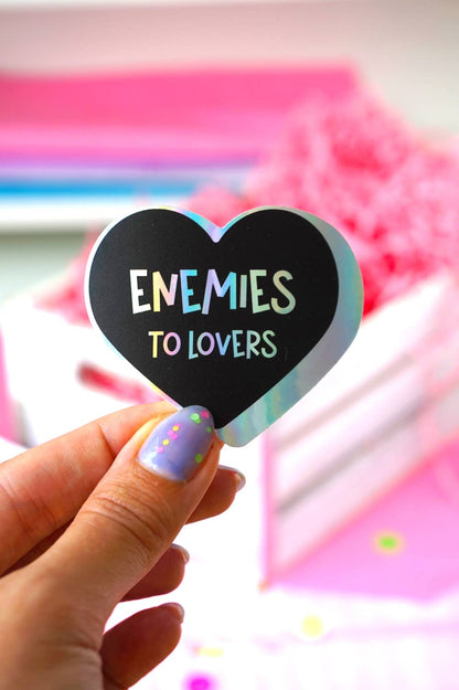 A hand with lavender glitter nail polish holds a heart-shaped black sticker that reads &