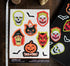 A sheet of vintage-style Halloween vinyl stickers displayed on a dark wooden surface next to a ruler and a black sticker roll. The stickers feature classic spooky faces and motifs including a skull, clown, witch, devil, cat in a hat, jack-o&