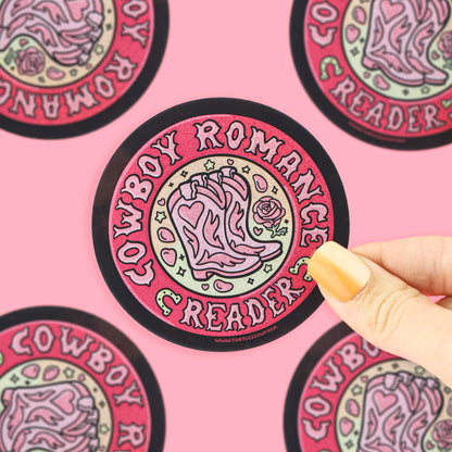 A hand with yellow nails holds a round sticker featuring a pink cowboy boot and the words &