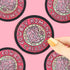 A hand with yellow nails holds a round sticker featuring a pink cowboy boot and the words &