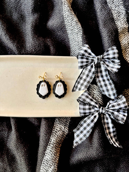 Black and white ghost-themed drop earrings displayed on a cream-colored rectangular tray, next to two black and white gingham ribbon bows on a dark textured fabric background.