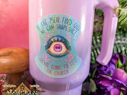 Pink mug with a colorful label featuring an eye design and text, surrounded by flowers and a pink background.