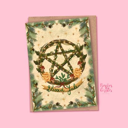A square greeting card with a kraft envelope, featuring a green pentagram wreath, pine branches, and the words &