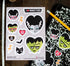 A sheet of Halloween-themed vinyl stickers featuring cartoon monsters, a cat, lightning bolts, and hearts, displayed on a dark wood surface next to a ruler and a black-and-white patterned phone case with matching stickers.