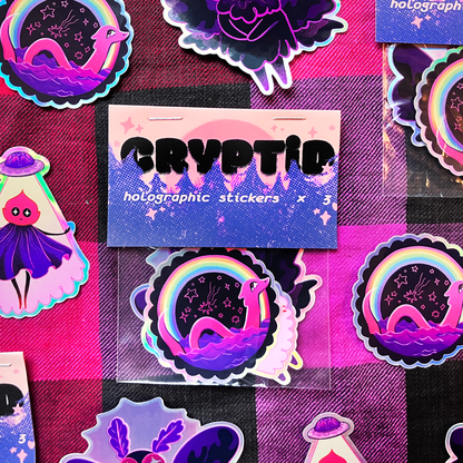 A set of colorful holographic stickers featuring a purple snake in a starry night sky under a rainbow, displayed on a pink and black checkered background. A central card reads &