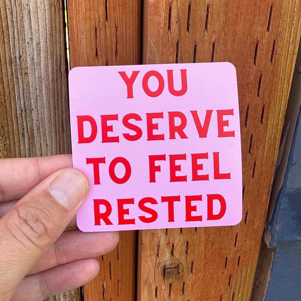 A hand holds a square pink sticker with bold red text that reads &