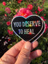 A hand holds a heart-shaped black sticker with holographic glitter edging and the text &