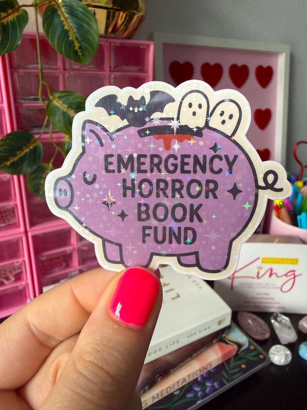 A hand with bright pink nail polish holds a die-cut sticker shaped like a purple pig. The sticker has black text reading &
