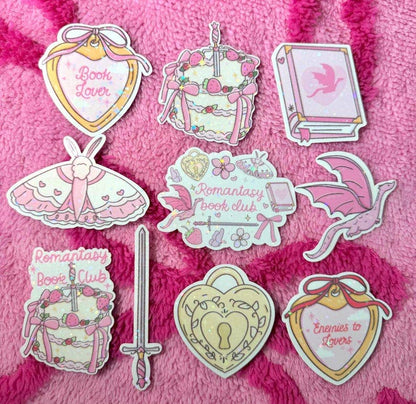 A set of cute, book-and-moth-themed enamel pins arranged on a pink textured fleece background. The pins feature illustrations of pink and cream-colored moths, a sword, a keyhole, and hearts with text such as &
