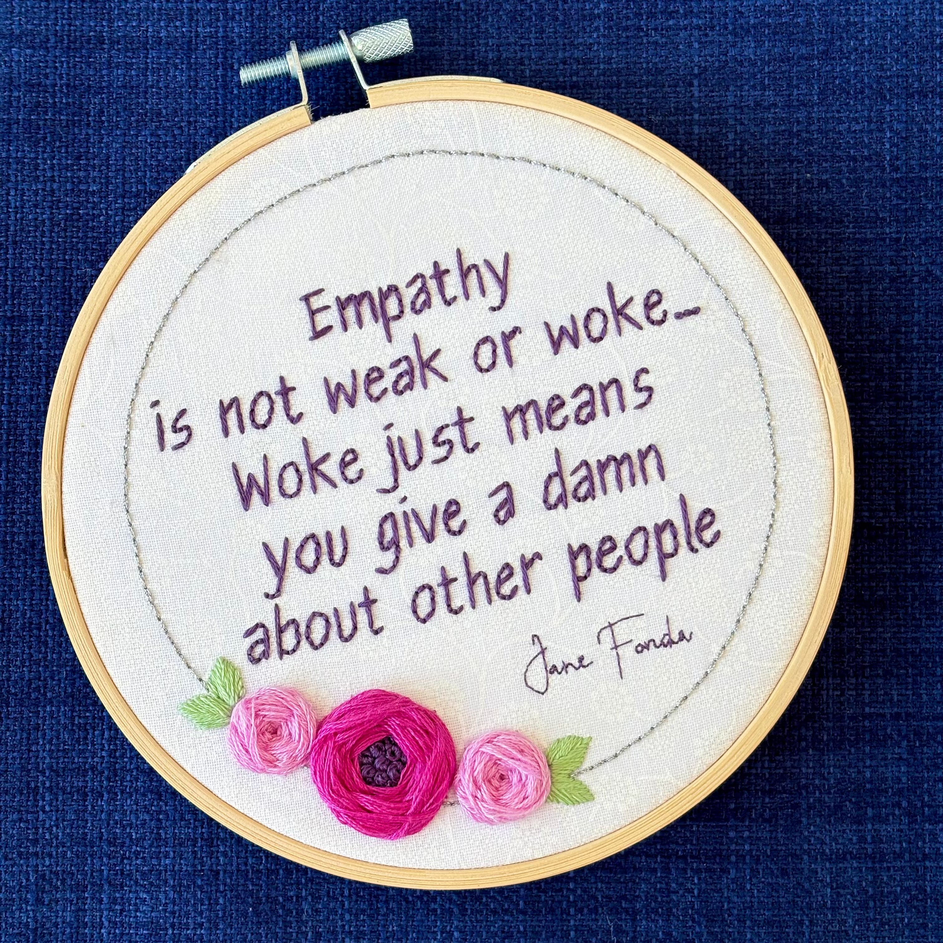 "Empathy is not weak or work...Woke just means you give a damn about other people. Jane Fonda." Quote hand embroidered in dark purple. Framed in silver circle floral wreath with bright fuchsia and pink roses and light green leaves. Hand stitched on white-on-white cotton. Displayed as fan wall art in light wood embroidery hoop.