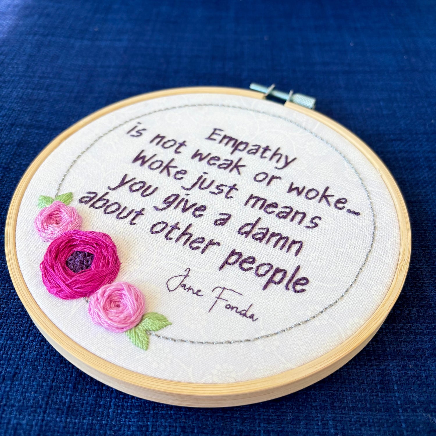 "Empathy is not weak or work...Woke just means you give a damn about other people. Jane Fonda." Quote hand embroidered in dark purple. Framed in silver circle floral wreath with bright fuchsia and pink roses and light green leaves. Hand stitched on white-on-white cotton. Displayed as fan wall art in light wood embroidery hoop.