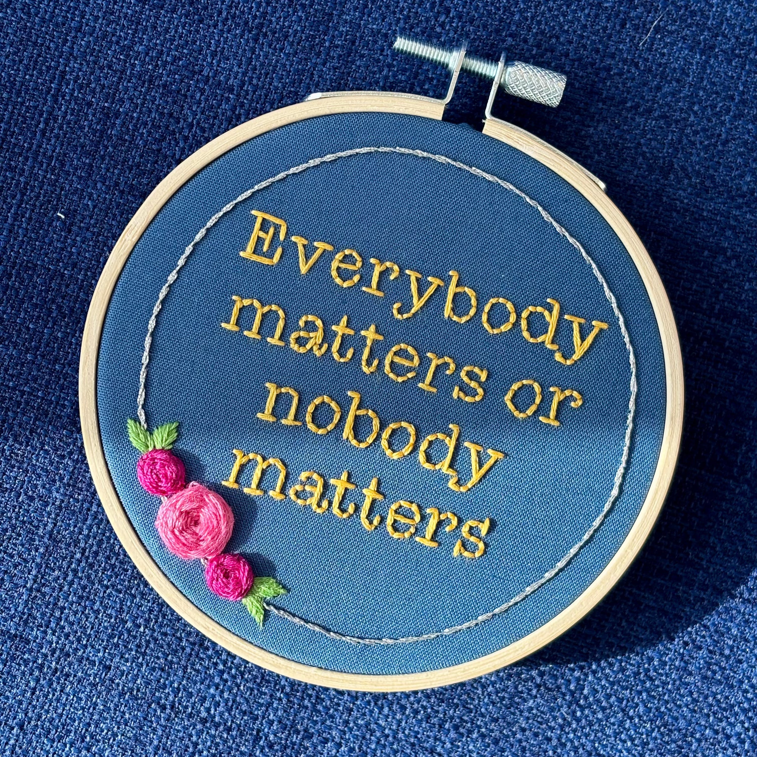 "Everybody matters or nobody matters." Quote hand embroidered in sunny yellow thread in a serif typewriter font. Framed with a ring of metallic silver thread, spring green leaves, and pink and fuchsia roses. Stitched on blue cotton fabric and displayed in a light wooden embroidery hoop as wall art.