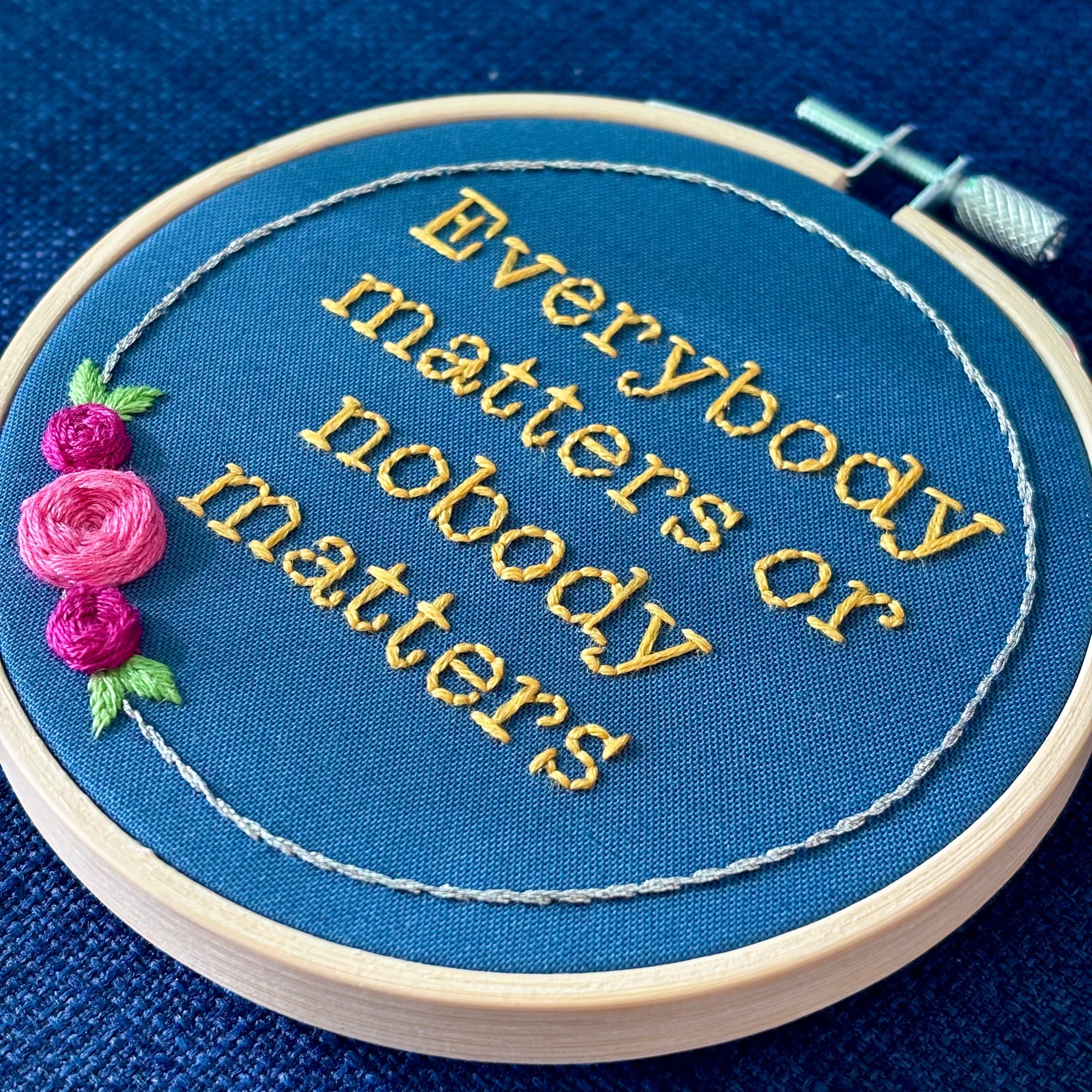 "Everybody matters or nobody matters." Quote hand embroidered in sunny yellow thread in a serif typewriter font. Framed with a ring of metallic silver thread, spring green leaves, and pink and fuchsia roses. Stitched on blue cotton fabric and displayed in a light wooden embroidery hoop as wall art.