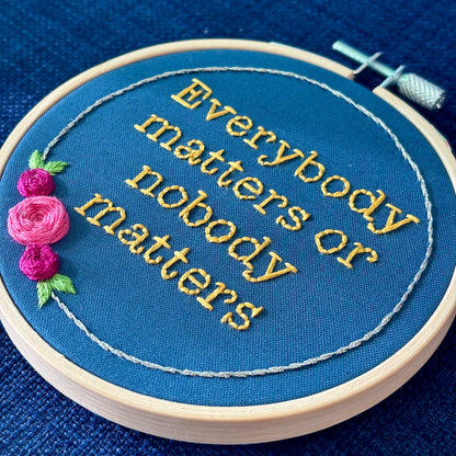 "Everybody matters or nobody matters." Quote hand embroidered in sunny yellow thread in a serif typewriter font. Framed with a ring of metallic silver thread, spring green leaves, and pink and fuchsia roses. Stitched on blue cotton fabric and displayed in a light wooden embroidery hoop as wall art.