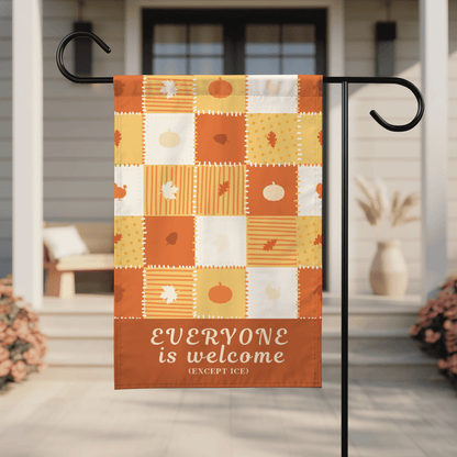 An autumn-themed garden flag hanging from a black metal stand on a front porch. The flag has a patchwork design in orange, yellow, and white with fall motifs like pumpkins, leaves, and stripes, and a bottom banner that reads &