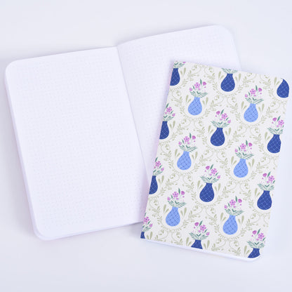 small pocket notebook with a pattern of blue vases and purple flowers on a sage green background, resting on an open notebook with dot grid pages