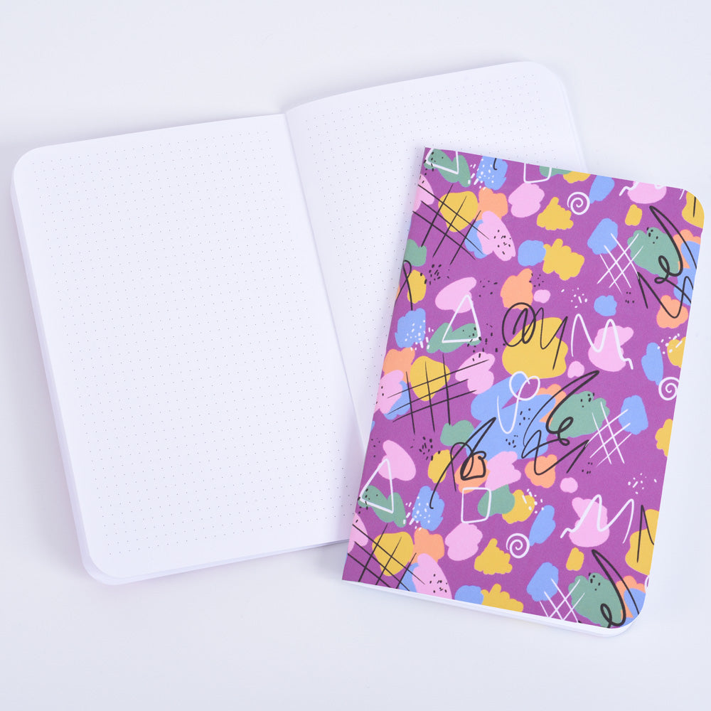 a small pocket notebook featuring a magenta abstract design with doodles and paint splotches resting on an open notebook with small dot grid paper