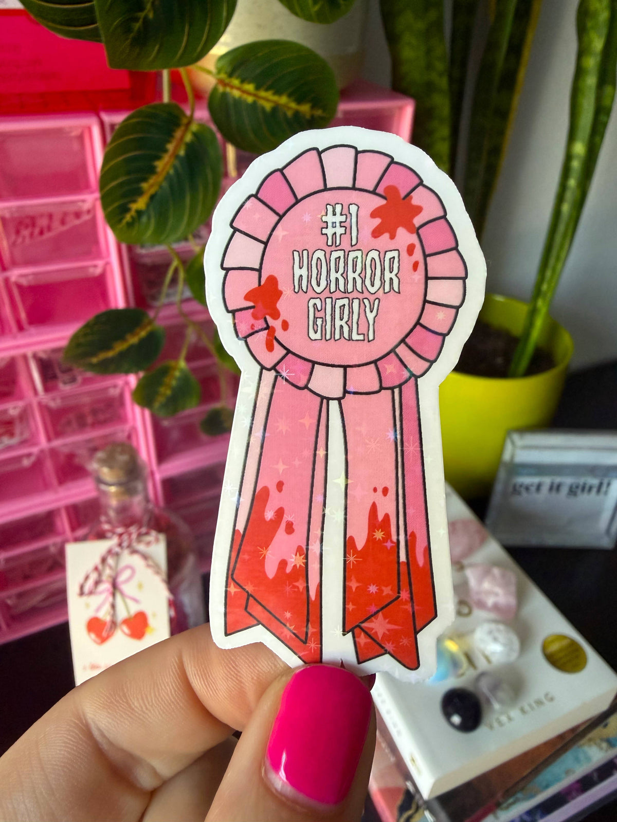 A hand with bright pink fingernails holds a die-cut sticker shaped like a pink ribbon award. The sticker reads “